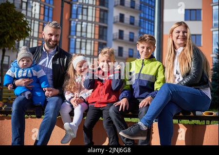 Zwei Erwachsene verbringen Zeit mit niedlichen Kindern und lächeln am sonnigen Frühlingstag. Vorderansicht der Eltern mittleren Alters, die mit vier entzückenden Kindern auf der Bank sitzen und sich im Stadtzentrum ausruhen. Familienkonzept. Stockfoto