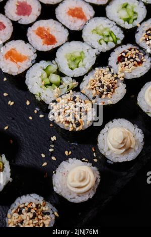 Draufsicht Maki-Brötchen mit Lachs, Thunfisch, Gurkensesame und Frischkäse liegen auf einem Sushi-Brett aus schwarzem Stein. Traditionelle japanische Küche. Stockfoto