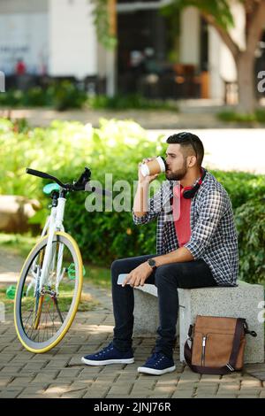 Selbstbewusster bärtiger Radler, der auf der Parkbank sitzt und duftenden Kaffee genießt, während er eine kurze Pause macht, ein Porträt in voller Länge Stockfoto