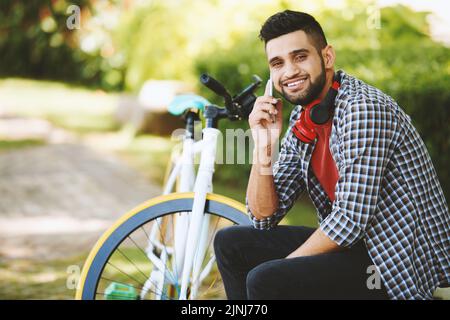 Fröhlicher indischer Mann, der mit toothy Lächeln auf die Kamera schaut, während er mit einem Freund auf dem Smartphone spricht und auf einer Parkbank sitzt, modernes Fahrrad im Hintergrund Stockfoto