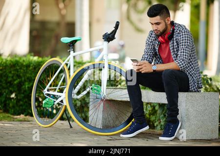 In voller Länge Porträt eines hübschen jungen Mannes, der mit einem Freund auf dem Smartphone schreibt, während er nach einer intensiven Radtour auf der Parkbank sitzt Stockfoto