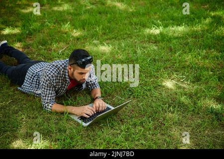 Gutaussehender bärtiger Designer in Casualwear, der auf grünem Rasen liegt, während er mit Hilfe eines Laptops an einem Start-up-Projekt arbeitete, Portraitaufnahme Stockfoto