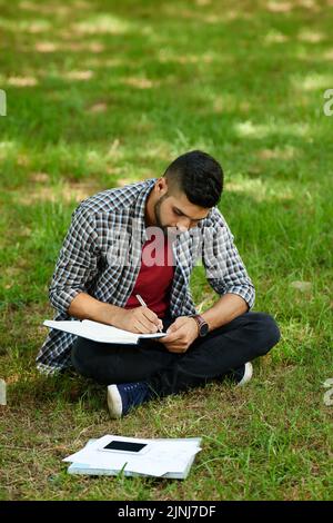 Konzentrierter indischer Student in Casualwear, der in Lotusposition auf grünem Rasen sitzt und sich während der Vorbereitung auf die Hochschulprüfung Notizen gemacht hat Stockfoto