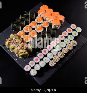 Auswahl an verschiedenen Arten von Sushi-Rollen auf schwarzem Steinbrett platziert. Sushi-Set mit Lachs, Gurken, Frischkäse, Avocado und Sesam. So Stockfoto