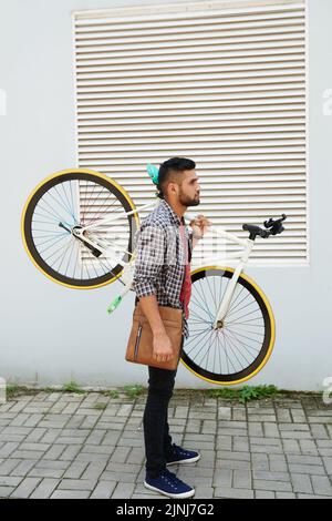 Profilansicht eines bärtigen jungen Mannes in Casualwear, der ein modernes Fahrrad auf der Schulter hält, während er an der Gebäudefassade steht, Ganzkörperportrait Stockfoto