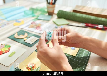 Nicht erkennbare junge Frau, die dekorative Elemente für ihre handgefertigte Grußkarte anfertigte, während sie am Holztisch saß Stockfoto