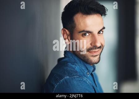 Porträt eines selbstbewussten und lässigen Mannes, der sich an einer grauen Wand lehnt und glücklich, stolz und froh aussieht, während er lächelt. Das Gesicht von stylisch, hübsch und jung Stockfoto