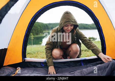 Porträtaufnahme eines attraktiven asiatischen Wanderers in Windjacke, der auf Geisterhacken sitzt und Zelt aufstellt, malerischer Blick im Hintergrund Stockfoto