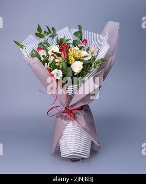 Blumenstrauß, der in helles Blumenpapier von zwei Arten gehüllt ist, steht auf blauem Hintergrund. Bouquet von weißen Rosen, farbigen Iris und roten Tulpen Stockfoto