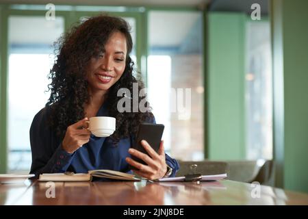 Lächelnde asiatische Geschäftsfrau, die in einem gemütlichen kleinen Café sitzt, duftenden Kaffee trinkt und mit einem Freund auf dem Smartphone schreibt Stockfoto