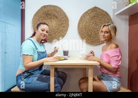 Junge Frauen blicken auf die Kamera und lächeln beim gemeinsamen Frühstück in einem Restaurant. Freundschafts- und Essenskonzept. Stockfoto