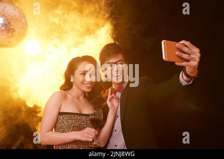Fröhliches asiatisches Paar, das Selfie auf dem Smartphone macht, während es die Party im Nachtclub genießt, hübsche Frau, die ein Glas Rotwein in der Hand hält Stockfoto