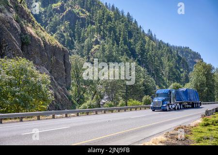 Leistungsstarker blauer großer Sattelschlepper, der schwere Ladung mit Plane auf einem flachen Sattelauflieger transportiert, der auf der Autobahn in Columbia Gorg unterwegs ist Stockfoto