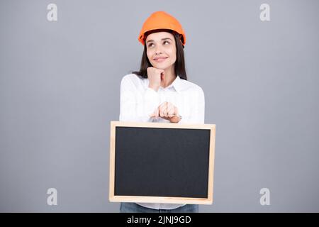 Junge lustige Frau Bauleiterin. Frau Baumeister isoliertes Porträt mit Schutzhelm und Brett. Grauer Hintergrund, leerer Speicherplatz, Kopierbereich. Stockfoto