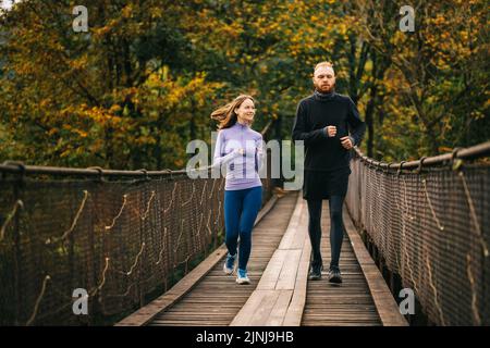 Junge Familie verbringt Zeit mit dem Nutzen. Gesundes, verliebtes Sportpaar läuft auf einer hölzernen Hängebrücke vor dem Hintergrund des Waldes zusammen Stockfoto