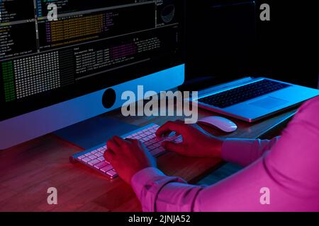 Nahaufnahme eines nicht erkennbaren Hackers, der vertrauliche Daten raubt, während er vor einem modernen Computer in einem dunklen Raum sitzt Stockfoto