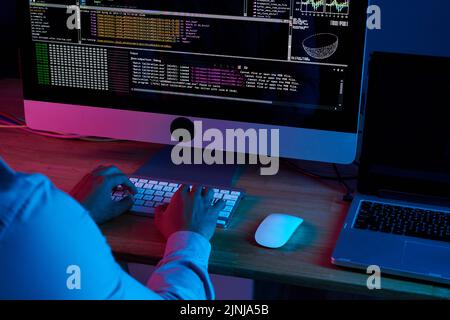 Nahaufnahme eines nicht erkennbaren Programmierers mit weißem Hemd, der vor einem modernen Computer sitzt und Programmiercode in einem dunklen Büro schreibt Stockfoto
