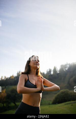 Eine junge Frau, die einen aktiven Lebensstil führt, führt Yoga-Übungen in einem offenen Berggebiet durch. Frau legt ihre Handflächen zusammen und hält ihr Gleichgewicht. Täglich t Stockfoto