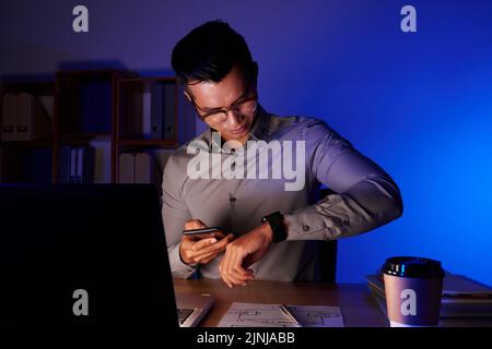 Porträt eines jungen asiatischen Coders, der die Armbanduhr anschaut, während er vor dem Computer sitzt und die Anzahl der Kollegen auf dem Smartphone anwählt, das Innere des dunklen Büros auf dem Hintergrund Stockfoto