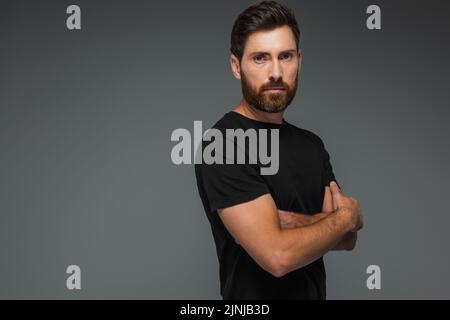 Porträt eines bärtigen Mannes in schwarzem T-Shirt, der die Kamera anschaut, während er mit gekreuzten Armen auf grauem, stockem Bild steht Stockfoto