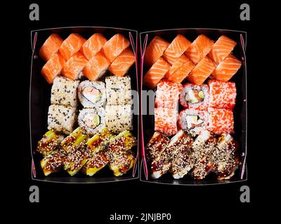 Draufsicht auf eine Auswahl an Sushi auf schwarzem Hintergrund in zwei Pappkartons, die zur Auslieferung bereit sind. Rainbow Sushi-Rollen, Uramaki, Hosomaki und Nigiri. Konzept Stockfoto