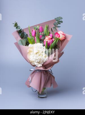 Handgefertigter Blumenstrauß zum Muttertag in einer festlichen Papierverpackung steht in einer Vase auf blauem Hintergrund. Wunderschön verpacktes Blumenarrangement mit Pink Stockfoto