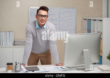 Porträt eines hübschen jungen Geschäftsmannes, der sich auf seinen Tisch lehnt und die Kamera anschaut Stockfoto