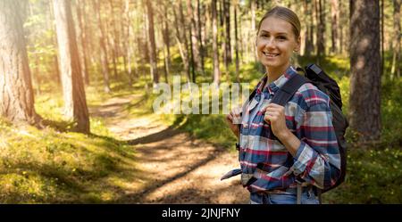 Lächelnde junge Frau im karierten Hemd mit Rucksack auf Waldweg stehen. Natur Abenteuer Wanderung. Copy space Stockfoto