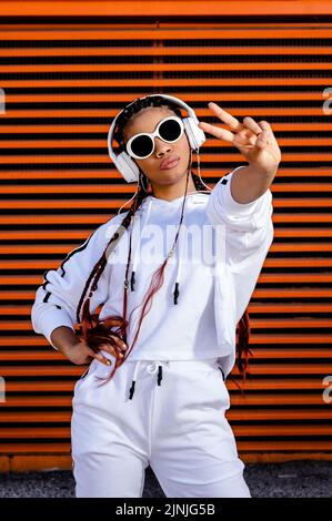 Eine junge afroamerikanische Frau mit geflochtenen Haaren, die urbane Sportkleidung mit Kopfhörern und Sonnenbrillen auf orangefarbenem Hintergrund trägt Stockfoto