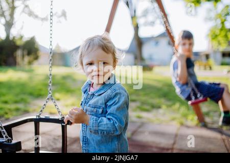 Kleine Geschwister spielen zusammen auf dem Spielplatz, wiegen sich auf einer Schaukel, genießen den Sommertag. Stockfoto