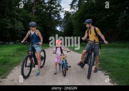 Junge Familie mit kleinem Kind, das im Sommer auf der Straße im Dorf Fahrräder reitet. Stockfoto