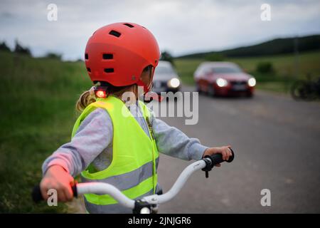 Rückansicht eines kleinen Mädchens in reflektierender Weste, das Fahrrad auf der Straße mit Autos hinter sich fährt, Konzept zur Aufklärung der Verkehrssicherheit. Stockfoto