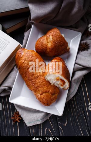 Hausgemachtes Gebäck. Frisch gebackene duftende Brötchen mit Marmelade im Inneren liegen neben einem offenen Buch auf einem Holztisch. Draufsicht auf ein ganzes Brötchen und ein aufgeschnittenes, auf w liegendes Brötchen Stockfoto