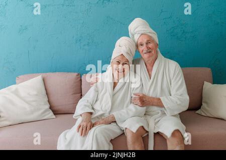Glückliches Seniorenpaar, das zusammen im Bademantel auf dem Sofa sitzt und eine schöne Zeit zu Hause hat. Stockfoto