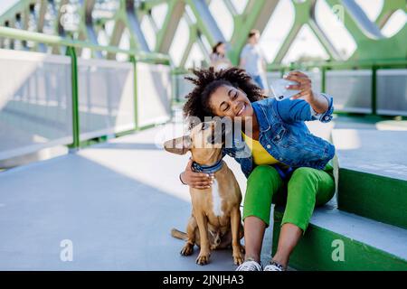 Multirassisches Mädchen, das mit ihrem Hund draußen auf der Brücke sitzt und sich ausruht, Selfie mit ihm macht und gemeinsam Freizeit verbringt. Konzept von Stockfoto