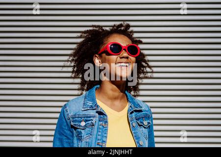 Porträt eines jungen glücklichen multirassischen Teenagers mit Sonnenbrille und Afro-Frisur, im Freien stehend. Stockfoto