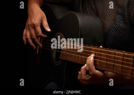 Nahaufnahme einer talentierten Frau, die in einem dunklen Raum sitzt und ihre Fähigkeiten beim Spielen von Akustikgitarre schärft Stockfoto