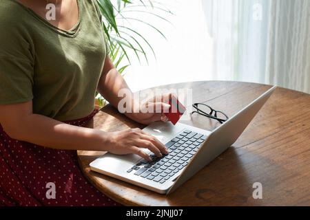 Nicht erkennbare Frau, die mit Kreditkarte in den Händen am Holztisch sitzt und in Nahaufnahmen beim Online-Shopping eingewickelt ist Stockfoto