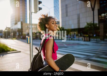 Multirassisches Teenager-Mädchen mit Rucksack und Skateboard, während des Sommers in der Stadt zu Fuß. Stockfoto