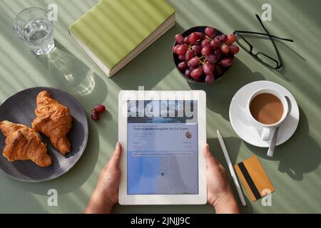 Aus dem hohen Winkel betrachten wir eine nicht erkennbare Frau, die ein köstliches Frühstück genießt, während sie am Küchentisch sitzt und ein digitales Tablet in Nahaufnahme verwendet Stockfoto