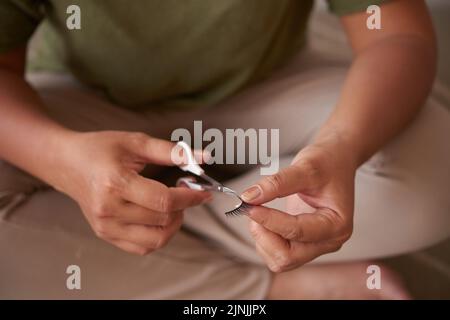 Unerkennbarer Make-up-Künstler, der in Lotusposition sitzt, während er mithilfe einer Schere die Länge falscher Wimpern kürzt, Nahaufnahme Stockfoto