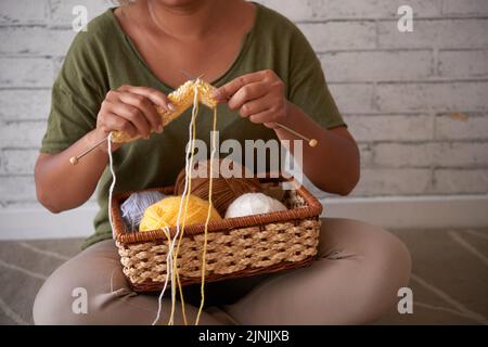 Frau, die in Lotusposition an der weißen Ziegelwand sitzt und in einen bunten Strickpullover eingewickelt ist, Nahaufnahme Stockfoto