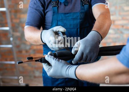 Zwei Erwachsene männliche Arbeiter in Overalls bereiten sich auf die Installation von Fußbodenheizungsrohren vor. Nahaufnahme der Hände von Reparaturwerkstätten in Arbeitshandschuhen, die Rohre befestigen und w Stockfoto