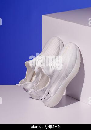 Zwei weiße Sneakers stehen auf einem weißen geometrischen Podium auf blauem Hintergrund. Die stylischen Damen-Sneakers stehen an einem Würfel angelehnt. Frühling und Sommer Mode c Stockfoto