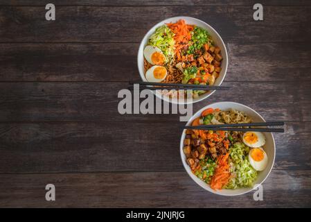 Variation von Ramen hausgemachte Suppe mit Nudeln, marinierten Tofu, gehackte Karotten, Zwiebeln, Salat und Brokkoli in zwei Schüsseln auf einem Holztisch. Stockfoto