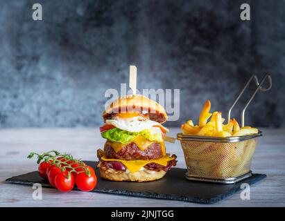 Leckerer hausgemachter Cheeseburger mit pommes auf schwarzem Schreibtisch auf grauem Hintergrund. Käse, Speck, Ei, Salat, Tomaten und Zwiebeln in hellem Brot. Unhea Stockfoto