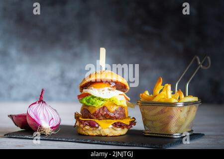 Leckerer hausgemachter Cheeseburger mit pommes auf schwarzem Schreibtisch auf grauem Hintergrund. Käse, Speck, Ei, Salat, Tomaten und Zwiebeln in hellem Brot. Unhea Stockfoto