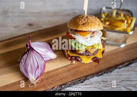 Hausgemachter Cheeseburger, der auf einem Holzschreibtisch mit Zwiebeln und pommes frites auf der Seite platziert wird. Grauer Hintergrund. Stockfoto