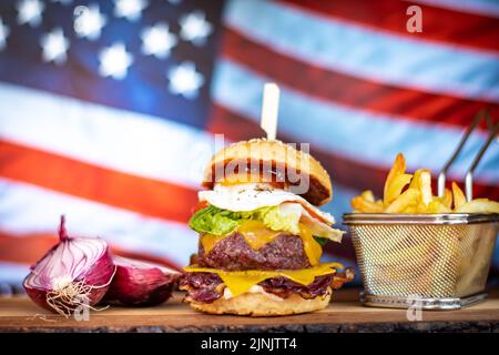 Cheeseburger mit pommes frites und Zwiebel wird auf einem Holzschreibtisch aufgestellt. Amerikanische Flagge im Hintergrund. Stockfoto