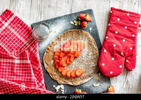 Hausgemachter Galette-Kuchen mit Erdbeeren und Mandel, auf schwarzem Schreibtisch und Holztisch platziert. Rotes Handtuch mit Kochwerkzeugen an der Seite Stockfoto